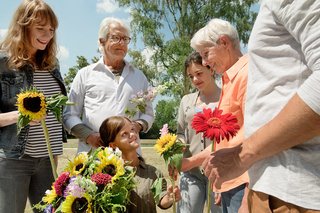 Unterschiedliche Menschen - alt und jung, Männer und Frauen, mit und ohne Behinderung - sind draußen und halten Blumen (unter anderem Sonnenblumen) in den Händen.
