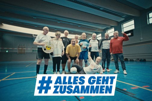 Gruppenfoto einer Volleyballmannschaft, die aus älteren Sportlern besteht, in einer Turnhalle. Darüber das Logo der Aktion Alles geht zusammen.
