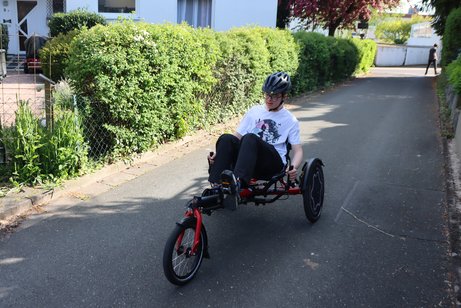 Balthasar unterwegs mit seinem Liegerad. Er trägt einen Fahrradhelm. Am Straßenrand steht seine Mutter und schaut ihm beim Fahren zu.
