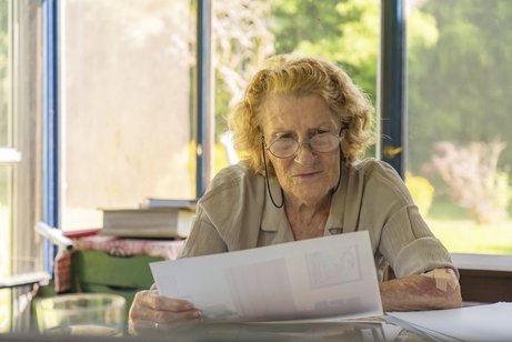 Konzentrierte ältere Frau mit Brille, die an ihrem Schreibtisch in einem sonnendurchfluteten Raum mit Blick ins Grüne sitzt und Papierkram durchgeht, möglicherweise Rechnungen oder medizinische Unterlagen