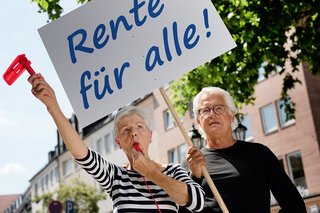 Eine ältere Frau und ein älterer Mann mit einer Demopappe mit der Aufschrift "Rente für alle!". Die Frau bläst in eine Trillerpfeife, hält eine Ratsche in der Hand. 