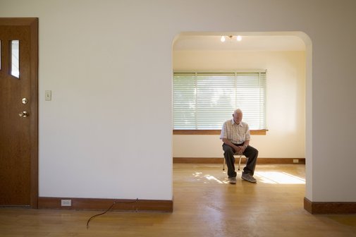 Blick in eine leerstehende Wohnung, in einem Zimmer sitzt allein ein alter Mann auf einem Stuhl.
