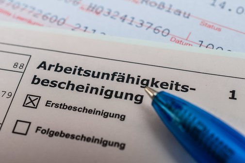 Arbeitsunfähigkeitsbescheinigung Eine Arbeitsunfähigkeitsbescheinung, auf der "Erstbescheinigung" angekreuzt ist, darauf liegt ein Kugelschreiber