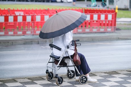 Eine Frau mit aufgespanntem Regenschirm sitzt auf ihrem Rollator auf einem Platz. Im Hintergrund ist ein Baustellenzaun zu sehen.