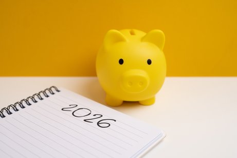 Ein gelbes Sparschwein steht auf einem wei&szlig;en Tisch, auf dem ein Notizbuch mit dem Eintrag "2026" liegt.