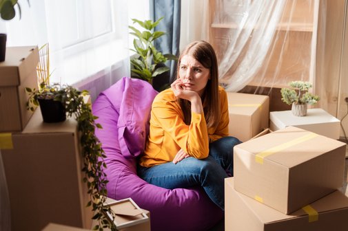 Haushalt auflösen Eine junge Frau sitzt auf einem Sitzsack in einer Wohnung zwischen Umzugskisten, sie blickt traurig und besorgt, hat das Kinn in die Hand gestützt.