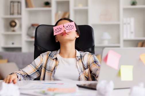 Eine junge Frau sitzt am Schreibtisch in einem B&uuml;ro, sie hat den Kopf zur&uuml;ckgelegt. Auf ihren geschlossenen Augen liegen Post-it-Zettel mit aufgemalten offenen Augen.