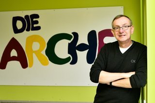 Bernd Siggelkow steht an eine Wand gelehnt, im Hintergrund sieht man ein großes Plakat mit der Aufschrift "Die Arche"