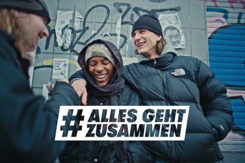 Drei Jugendliche stehen vor einer mit Graffiti besprühten Wand, sie lachen zusammen, einer hat den Arm über die Schulter des anderen gelegt. Darüber das Logo der Aktion Alles geht zusammen.