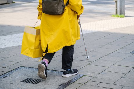 Person in gelben Regenmantel läuft mit einem Blindenstock entlang eines Blindenleitsystems 