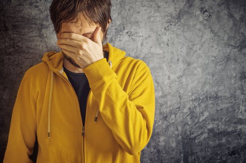 Ein Mann in gelber Sweatjacke steht vor einer grauen Wand, er hält sich eine Hand vor die Augen, senkt den Kopf