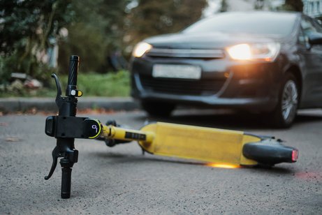 Ein gelber E-Scooter liegt umgest&uuml;rtzt auf der Stra&szlig;e. Ein Auto, dessen Scheinwerfer an sind, steht weniger Meter entfernt. 