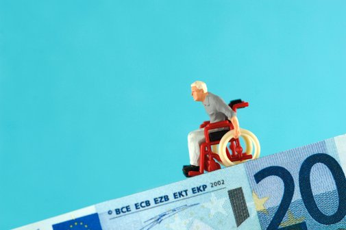 Symbolbild: Ein 20-Euro-Schein bildet eine absch&uuml;ssige Rampe, auf der eine Mini-Figur eines alten Mannes im Rollstuhl bergab rollt