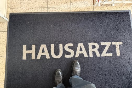 Jemand steht auf einer Fu&szlig;matte, auf der "Hausarzt" in Blockbuchstaben geschrieben steht