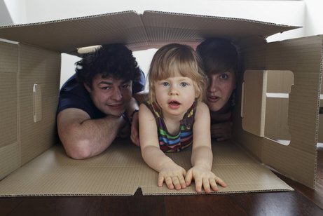 Wohnkostenlücke im Bürgergeld Eine junge Familie: Vater, Mutter und ein kleines Mädchen schauen gemeinsam aus einem großen Pappkarton heraus, der auf dem Boden liegt.