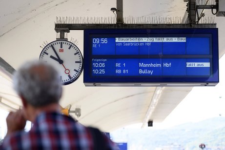 Rückenansicht eines älteren Mannes an einem Bahnstein am Bahnhof Koblenz, man sieht die digitale Anzeigetafel mit den angekündigten Zügen sowie eine Bahnhofsuhr.