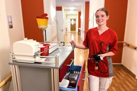 Das Foto zeigt Michelle. Sie steht neben einem Medikamentenwagen im Flur des Pflegeheimes und hält ein Blutdruckmessgerät in der Hand. 