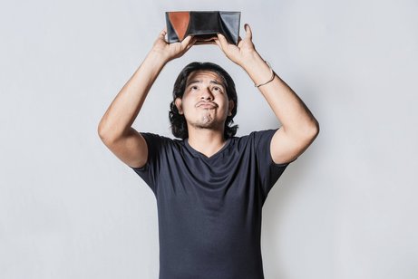 Ein Mann hält seine leere Brieftasche umgedreht über den Kopf, er schaut mit traurigem Gesichtsausdruck, ob etwas herausfällt.