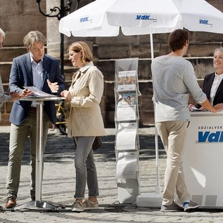 Ein VdK-Infostand in einer Fußgängerzone, eine Frau spricht mit einem Passanten, am Stehtisch daneben unterhalten sich Menschen. 
