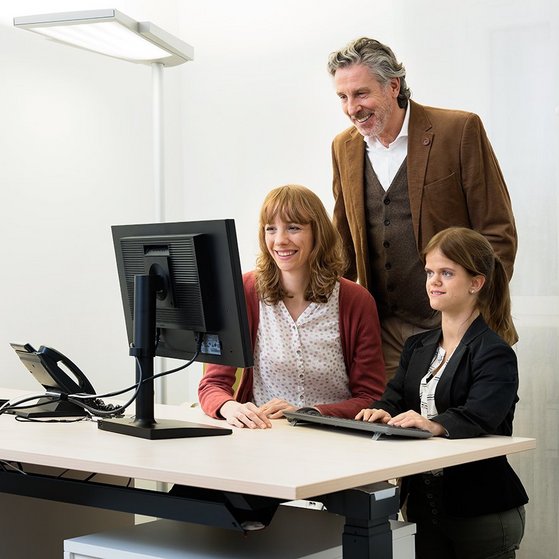Zwei Frauen und ein Mann stehen gemeinsam in einem Büro an einem Computer, blicken auf den Bildschirm