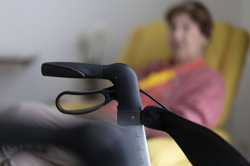 Seniorin sitzt in ihrem Zimmer im Seniorenheim hinter einem Rollator in einem gelben Sessel. Der Rollator im Bildvordergrund ist scharf zu sehen, die Seniorin im Hintergrund verschwommen.
