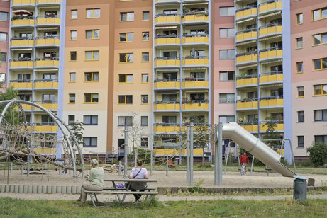 H&auml;userfassaden von gro&szlig;en Mehrfamilienh&auml;usern/Hochh&auml;usernm davor ein Spielplatz und eine Bank, auf der ein Mann und eine Frau sitzen.