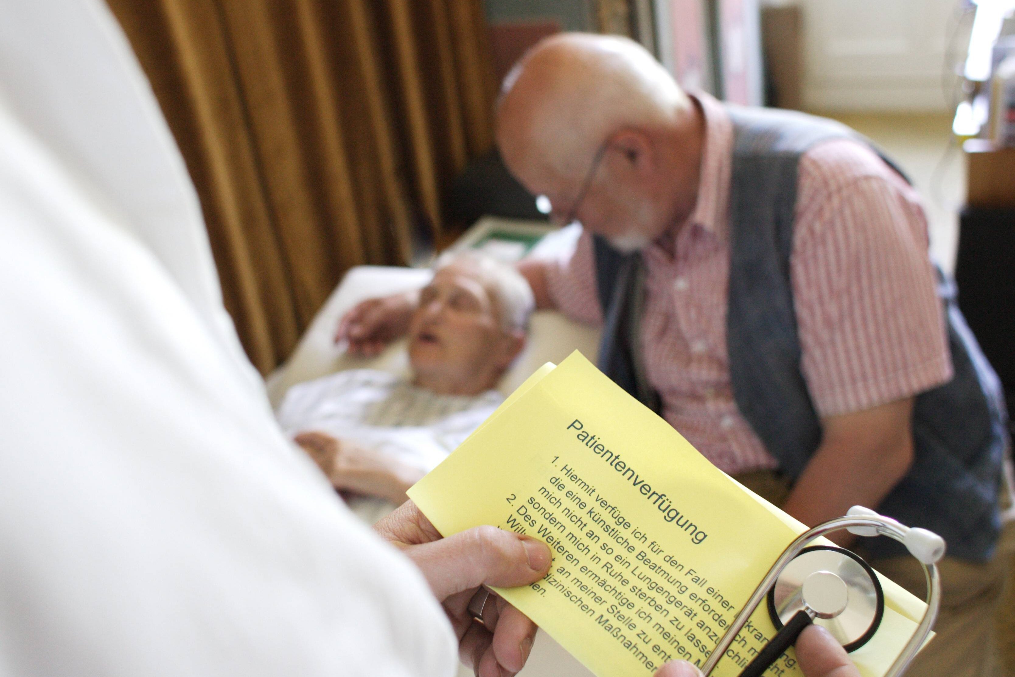 Patientenverfügung Eine alte Frau liegt im Bett, ein Arzt hält ihre Patientenverfügung in der Hand.