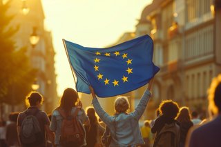Das Bild eine Demonstration mit einer Europafahne. Die Menschenmenge in einer Straße ist von hinten fotografiert. Die Sonne scheint von vorne und lässt die Fahne und die Menschen aufleuchten.