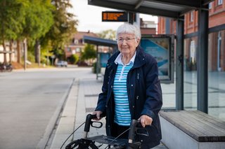 Eine ältere Frau wartet mit einem Rollator an einer Bushaltestelle.