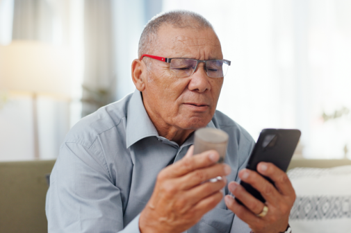 Ein Mann sitzt auf dem Sofa und checkt per Smartphone eine Medikamentendose.