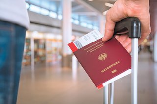 Eine Person, mit Rollkoffer und Reisepass in den Händen, hält sich in einem Flughafenterminal auf.