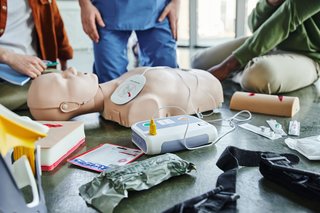 Das Bild zeigt eine CPR-Puppe, Defibrillator, Wundversorgungssimulatoren, Kompressionstourniquet, Bandagen und Spritzen in der Nähe von Ausbildern und Lernenden.