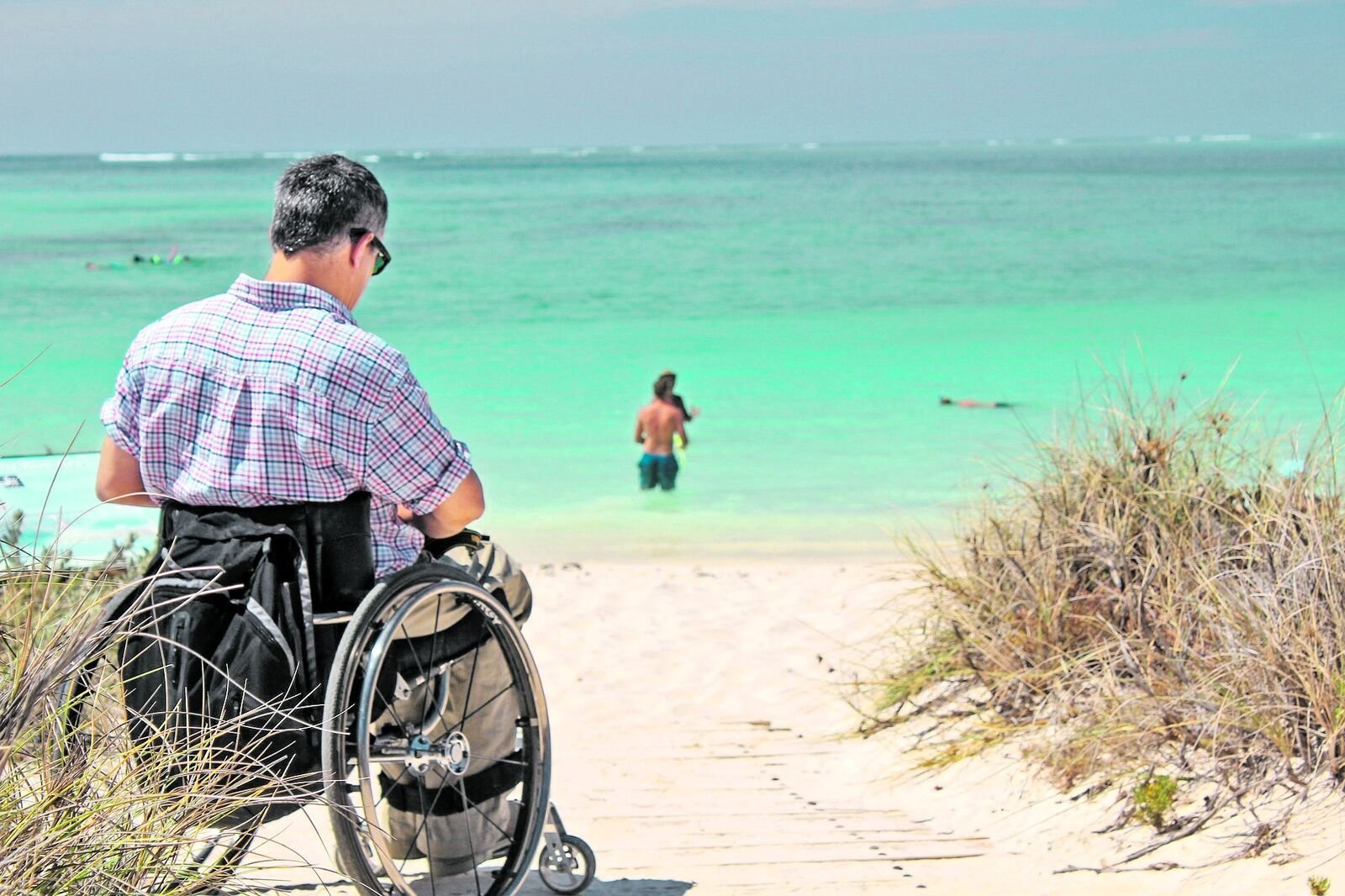 Eine Strandbild mit einem Mann im Rolli, viel Sand und Meer.