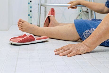 Das Bild zeigt eine gestürzte Frau. Die Frau sucht mit einer Hand halt am Gestell eines Badehockers.
