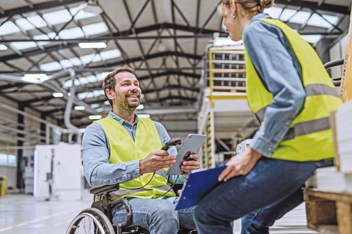 In einer Produktionshalle sind ein Mitarbeiter im Rollstuhl und eine Mitarbeiterin im Gespräch.