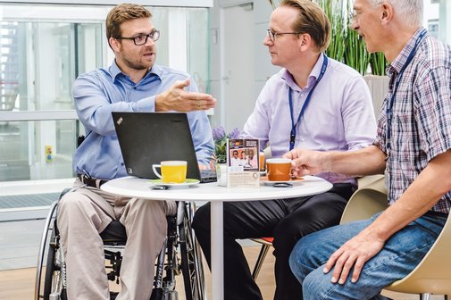 Drei Männer, davon einer im Rollstuhl, sitzen an einem Tisch und führen ein Gespräch.