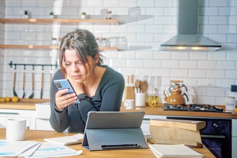 Eine junge Frau sitzt in der K&uuml;che und informiert sich per Smartphone.