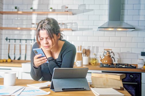 Eine junge Frau sitzt in der K&uuml;che und informiert sich per Smartphone.