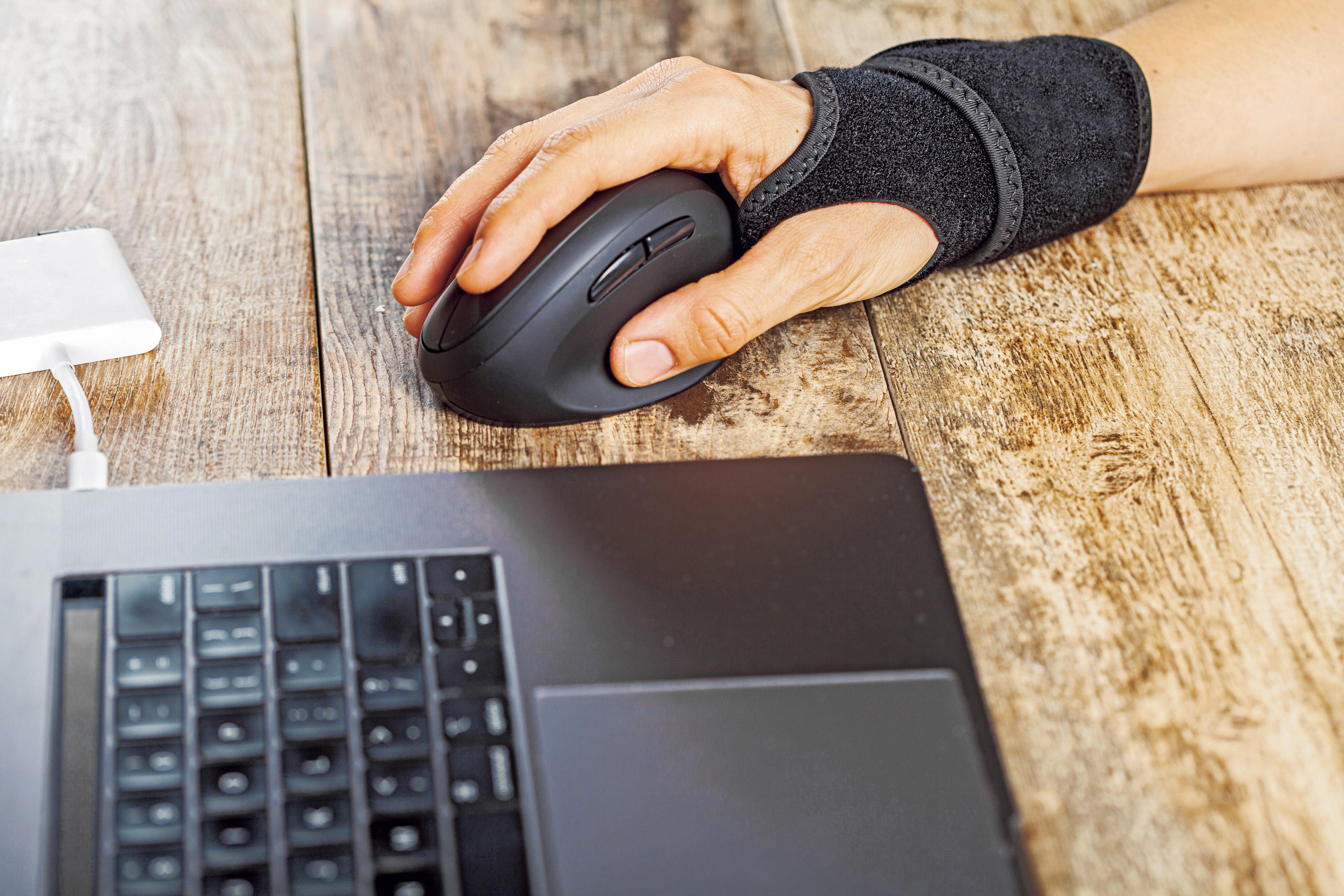 Das Bild zeigt ein Notebook und eine Hand mit Handgelenkmanschette und einer Spezialmaus die das ergonomische Arbeiten unterstützen. 