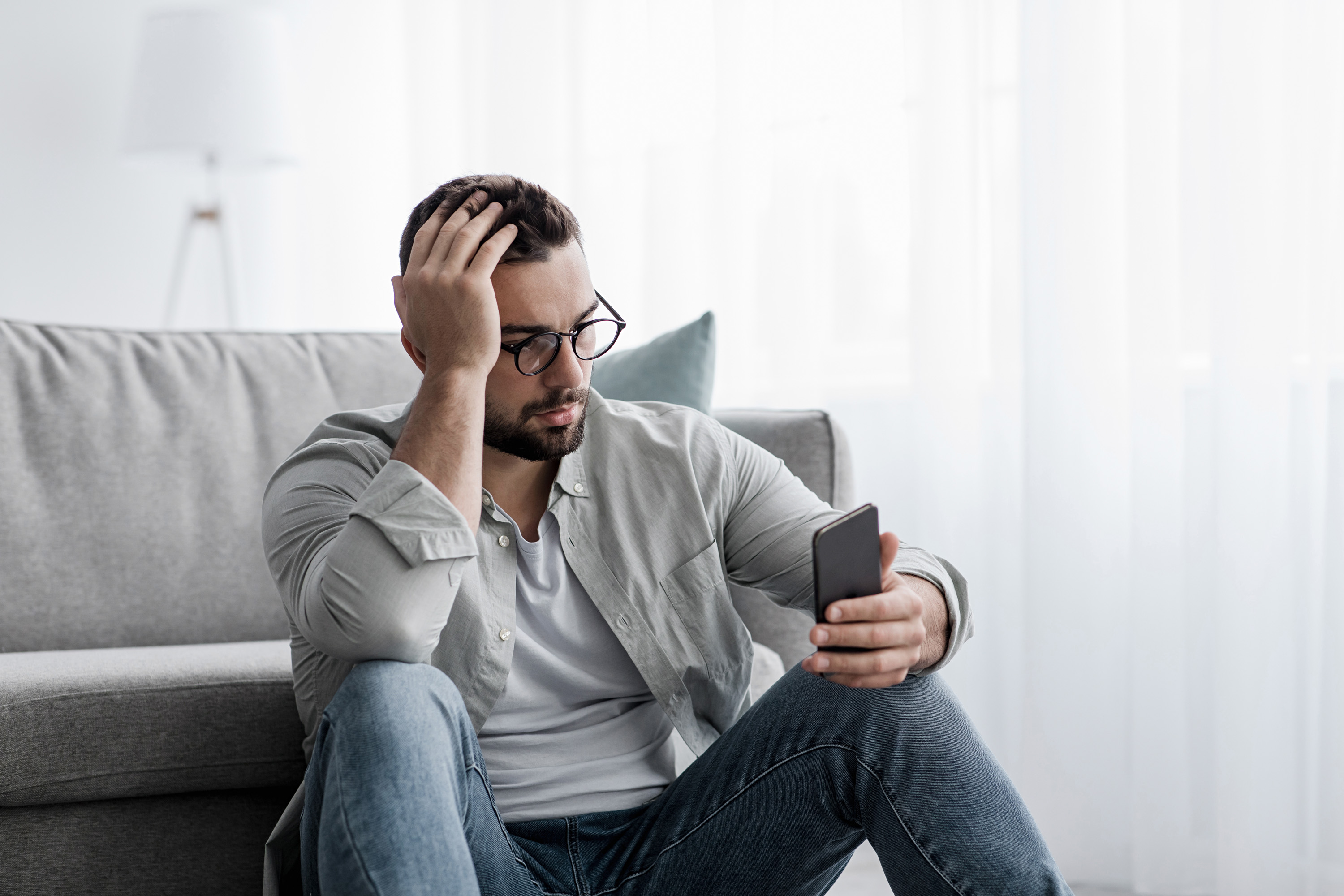 Eine auf dem Fußboden sitzende Person blickt nachdenklich auf sein Smartphone.